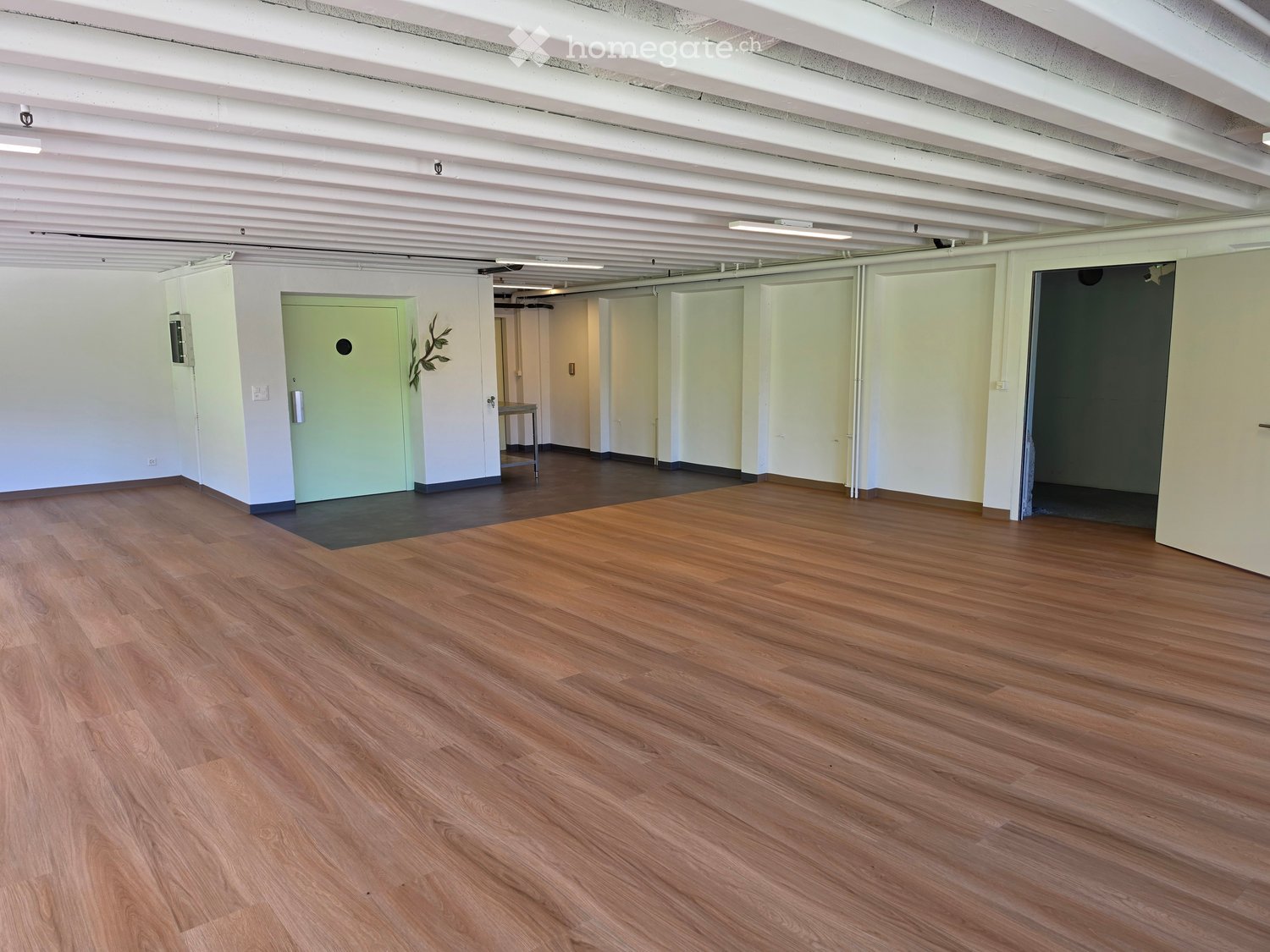 Empty room with wooden flooring, white walls, and several doors leading to other spaces. The room is well-lit with exposed ceiling beams.
