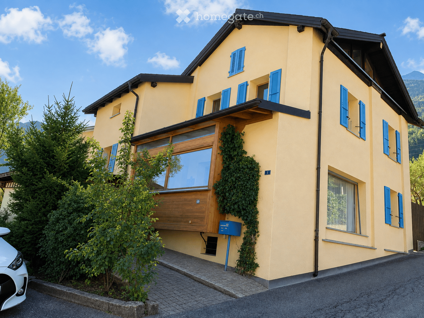 Two-story yellow house with blue shutters, wooden front door, and ivy-covered wall. The house features a balcony and is surrounded by greenery and a parked car.