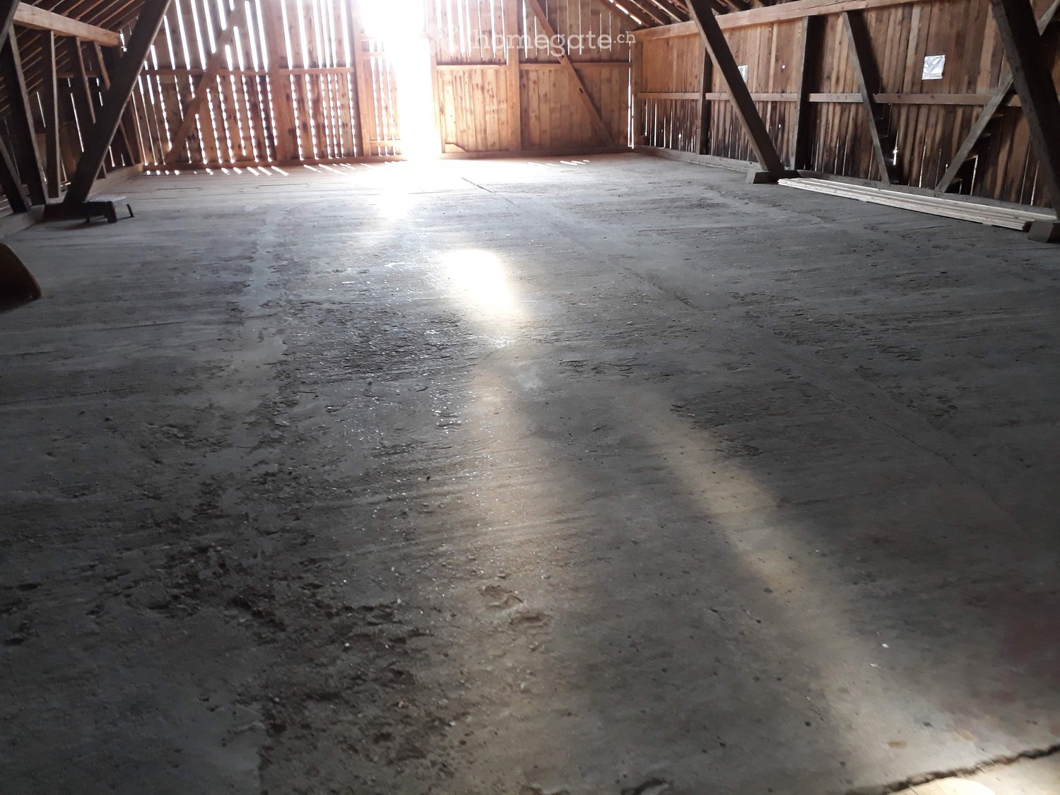 Empty barn interior with wooden beams, concrete floor, and natural light from windows.