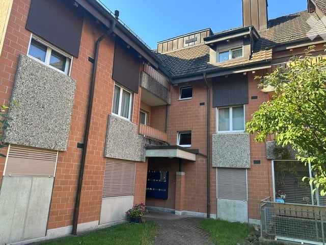 Multi-story brick apartment building with multiple windows, balconies, and a small garden area in front.