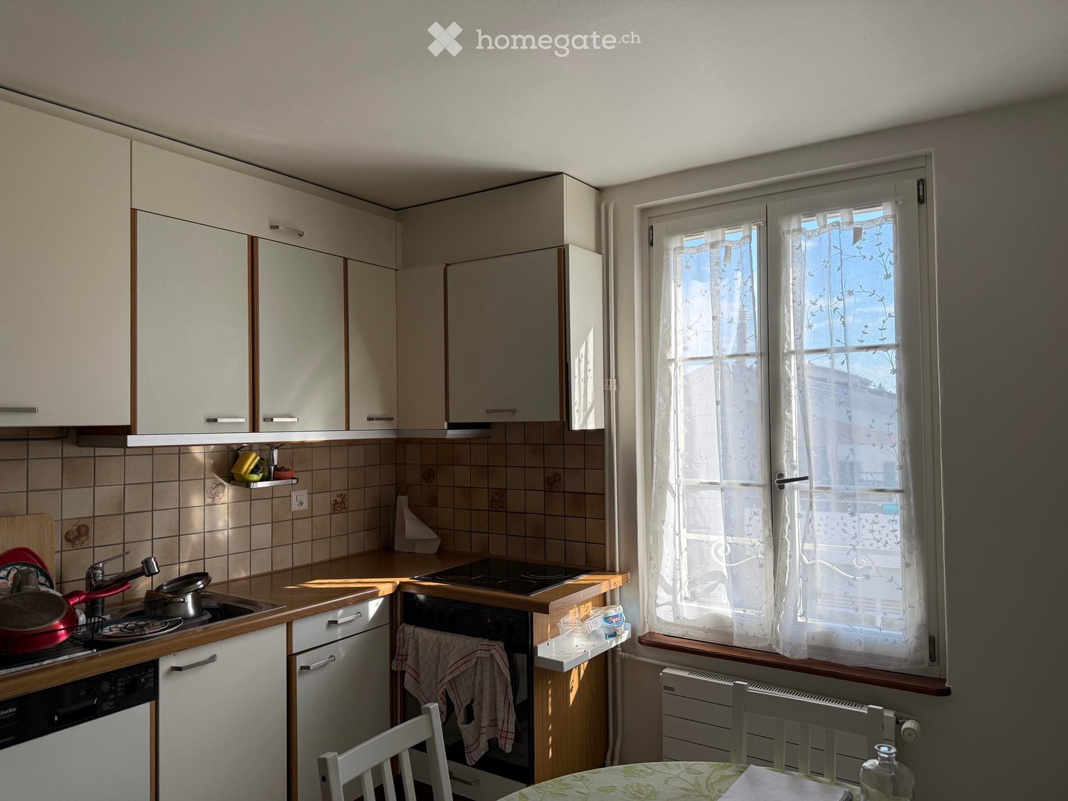 A small kitchen with white cabinets, a tiled backsplash, and a window with sheer curtains. The counter has a sink, stove, and various kitchen items, and there is a small dining area with a table and chairs.
