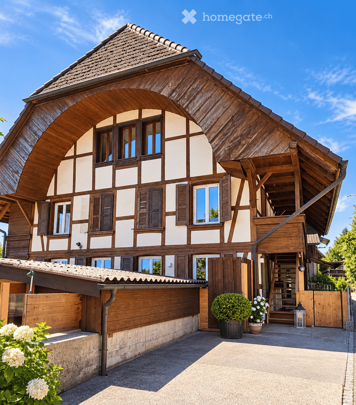 Traditional wooden house with a steeply pitched roof, white and brown facade, multiple windows with shutters, and a covered entrance area with steps leading up to the door.