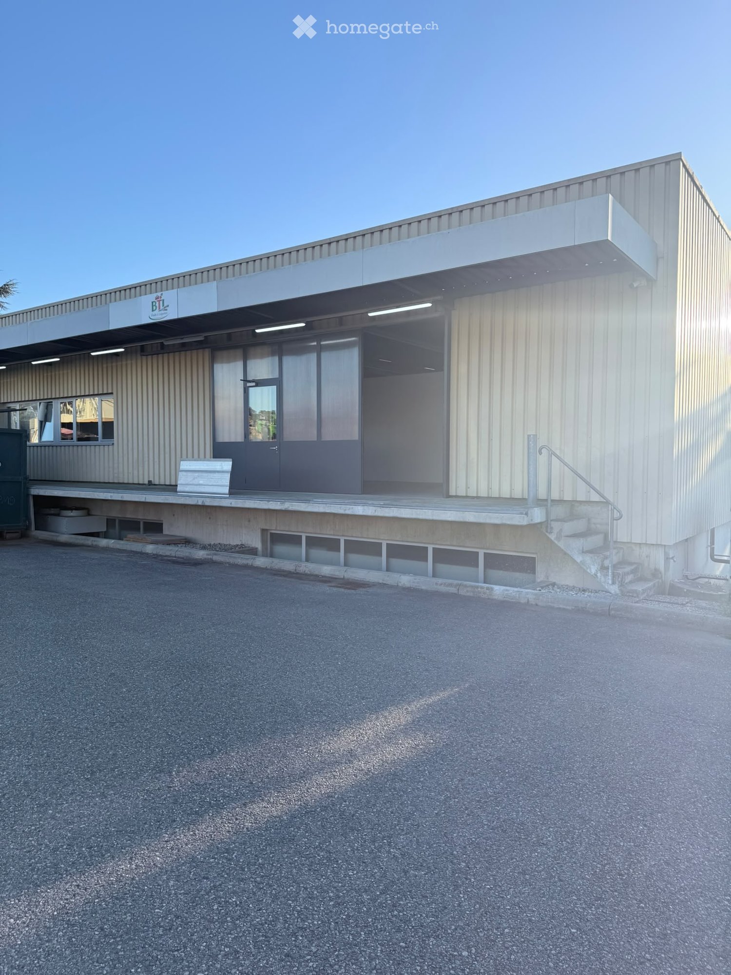 Modern industrial-style building with a metal facade, large windows, and a covered entrance area. Features a concrete staircase leading to the entrance and a small bench near the entrance.