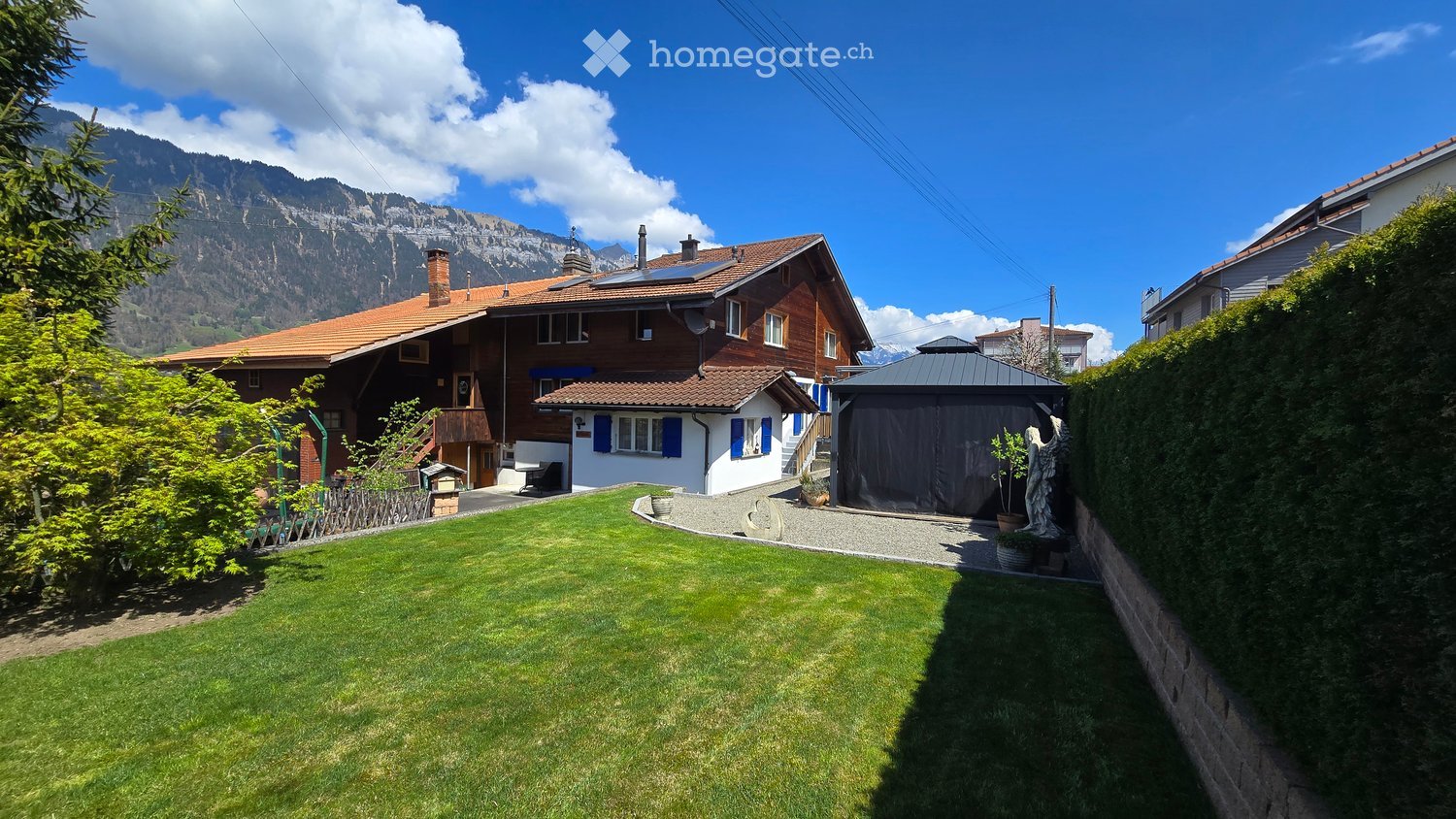 A well-maintained garden with a lawn, a small shed, and a statue, with a traditional house and a scenic mountain view in the background.