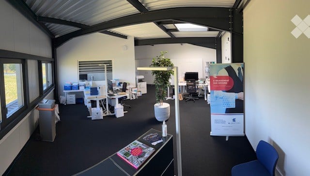 Modern office space with desks, chairs, computers, a potted plant, and a poster stand. The room features large windows and a high ceiling with exposed beams.