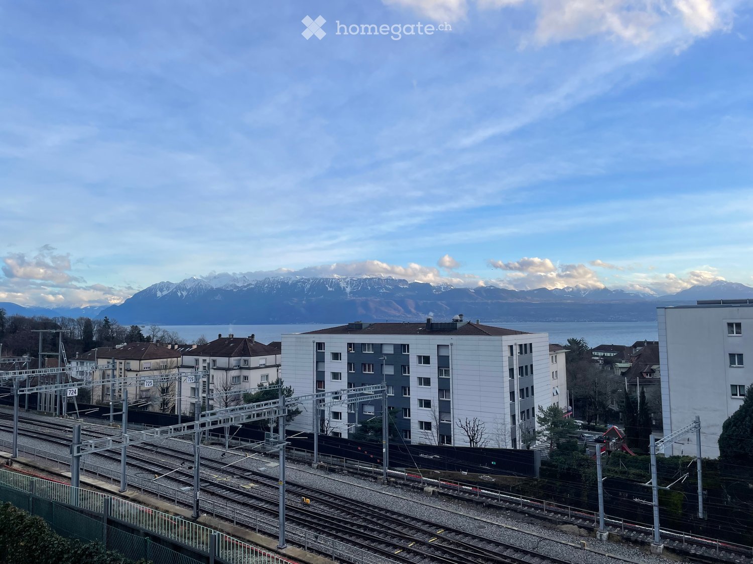 Modern multi-story residential building with white and grey facade, located near train tracks with a scenic mountain and lake view in the background.