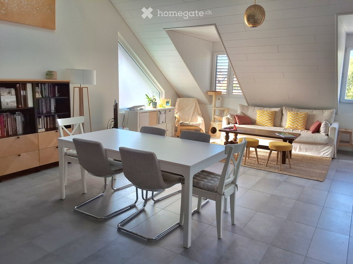Bright attic living space with a white dining table and chairs, a sofa with colorful cushions, a coffee table, a bookshelf, and a cat tree. The room features exposed wooden beams and large windows allowing natural light.