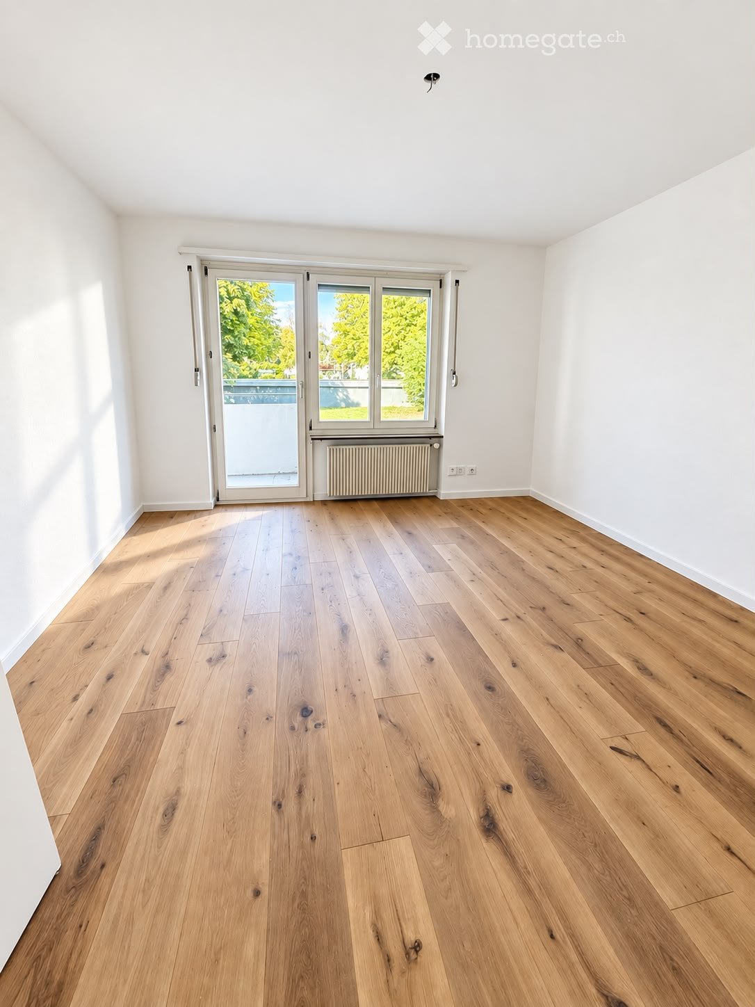 Empty room with light wooden flooring, white walls, and a large sliding glass door leading to a balcony or outdoor area.