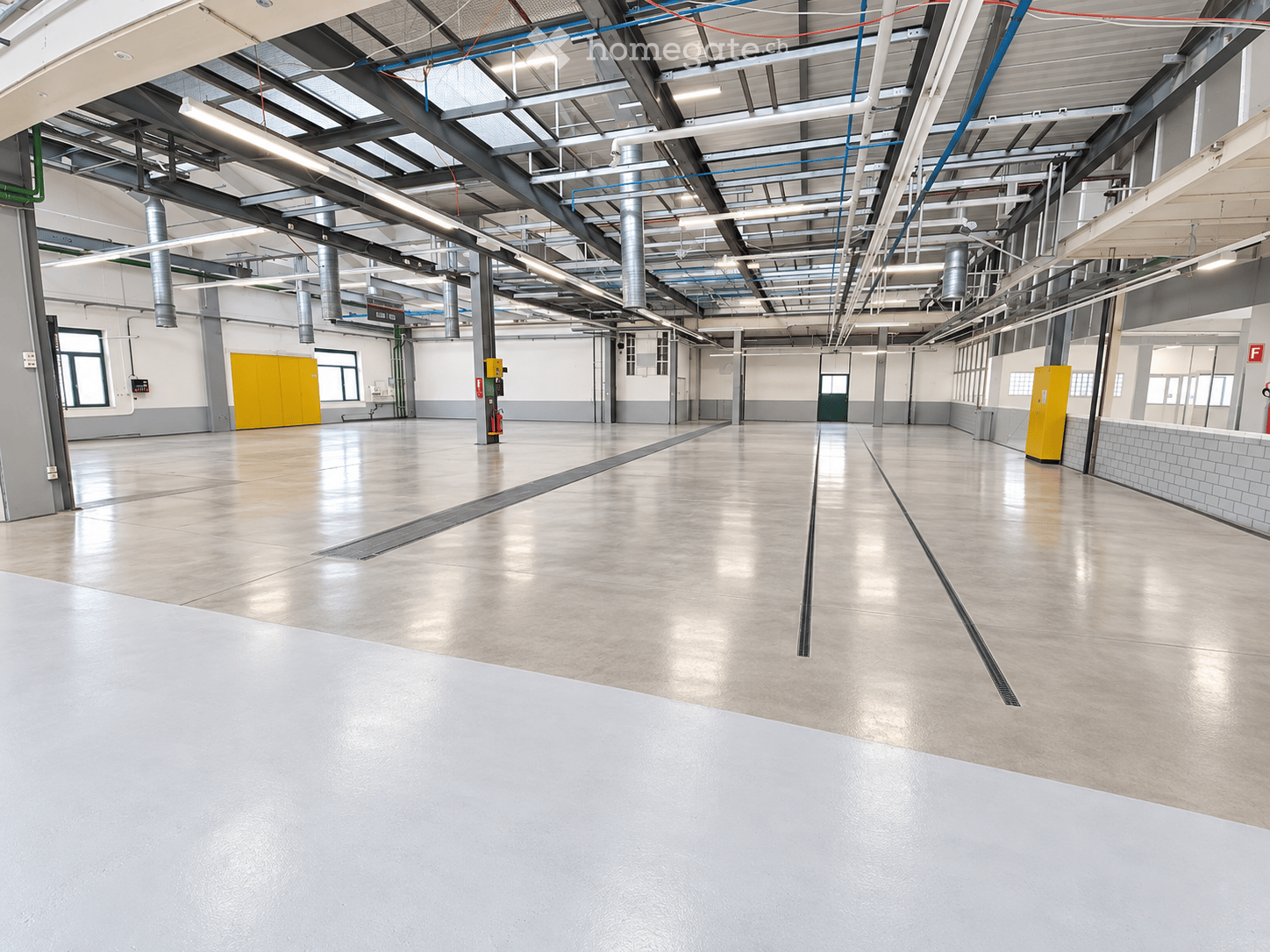 Large, empty industrial-style room with concrete floors, exposed metal beams, and large windows. No furniture or equipment is present.