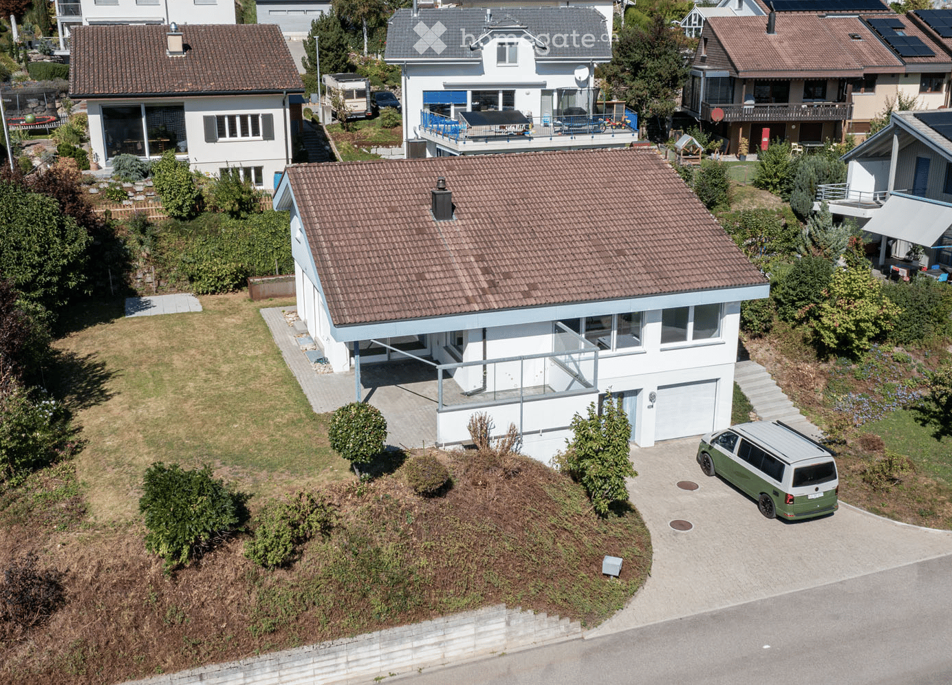 Aerial view of a modern two-story house with a tiled roof, white exterior, and a garage. The house features a raised ground floor, a balcony, and is surrounded by greenery and neighboring houses.
