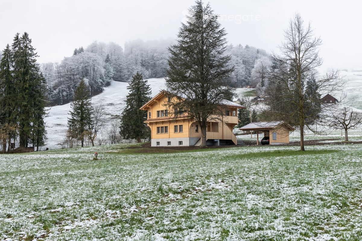 A two-story yellow house with a balcony and a small garage, surrounded by snow-covered trees and a field.