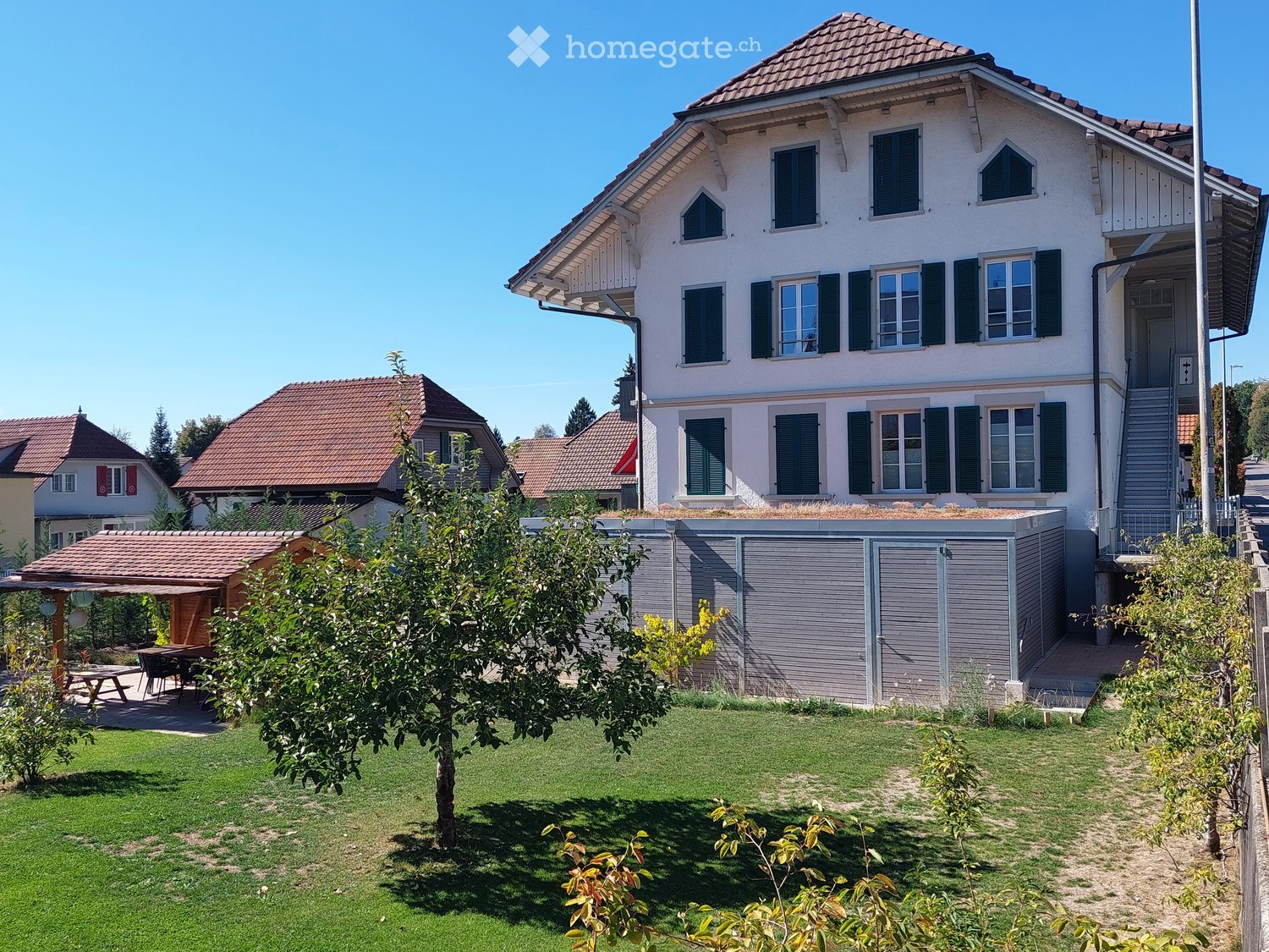 Two-story house with white walls, green shutters, and a red-tiled roof. The house features a modern garage with grey doors and a small patio area. The surrounding yard includes a well-maintained lawn, a tree, and a small outdoor seating area with a table