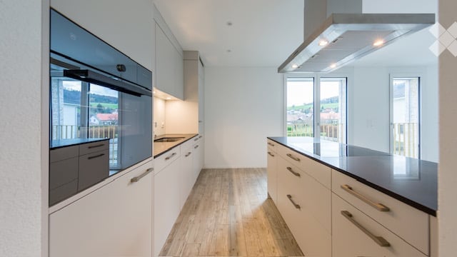 Modern kitchen with white cabinets, stainless steel oven, countertop, and wooden flooring. Large windows offer a scenic view.