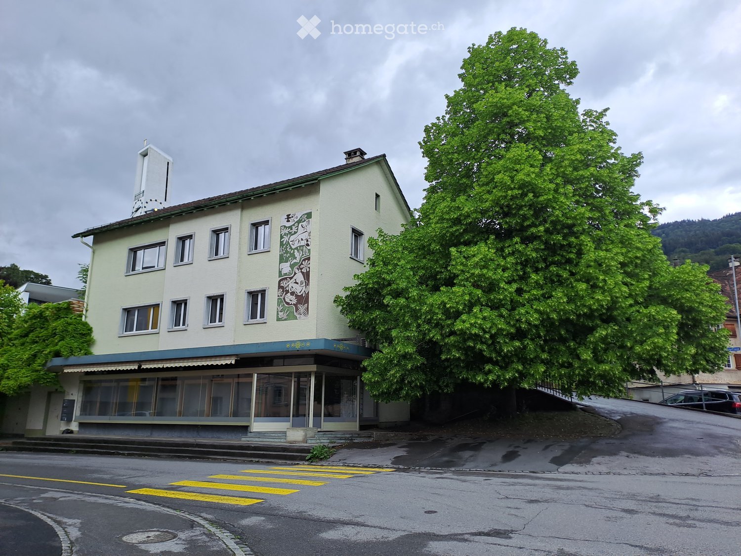 Two-story building with light yellow facade, multiple windows, and a mural on one side. The building has a small awning over the entrance and is surrounded by trees and a road with a crosswalk.