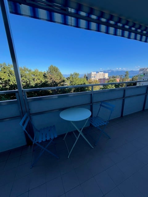 Balcony with a striped awning, a small round table, and two folding chairs, overlooking a scenic view of trees and a distant cityscape.