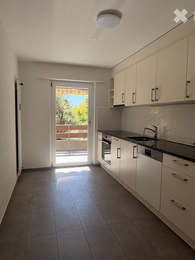 Modern kitchen with white cabinets, dark countertops, and a sliding glass door leading to a balcony. Features include a sink, faucet, oven, and dishwasher.
