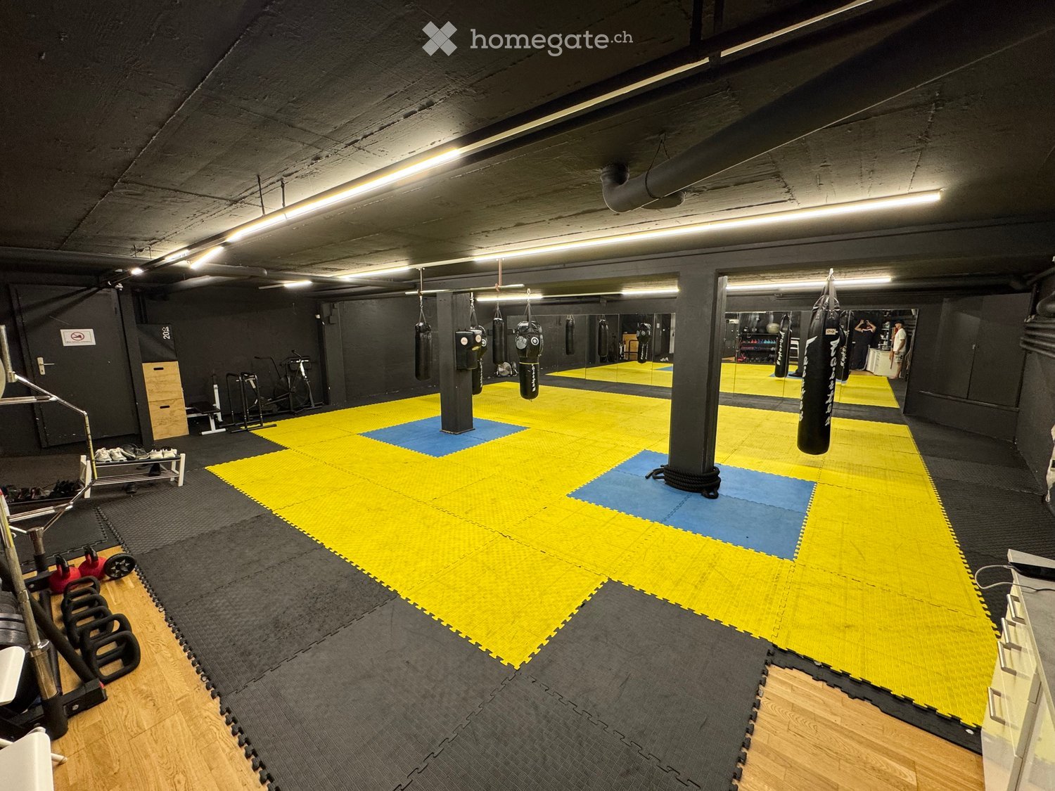 A spacious gym area with a large yellow and blue mat, boxing bags hanging from the ceiling, weights, a bench, and a mirror on the wall.