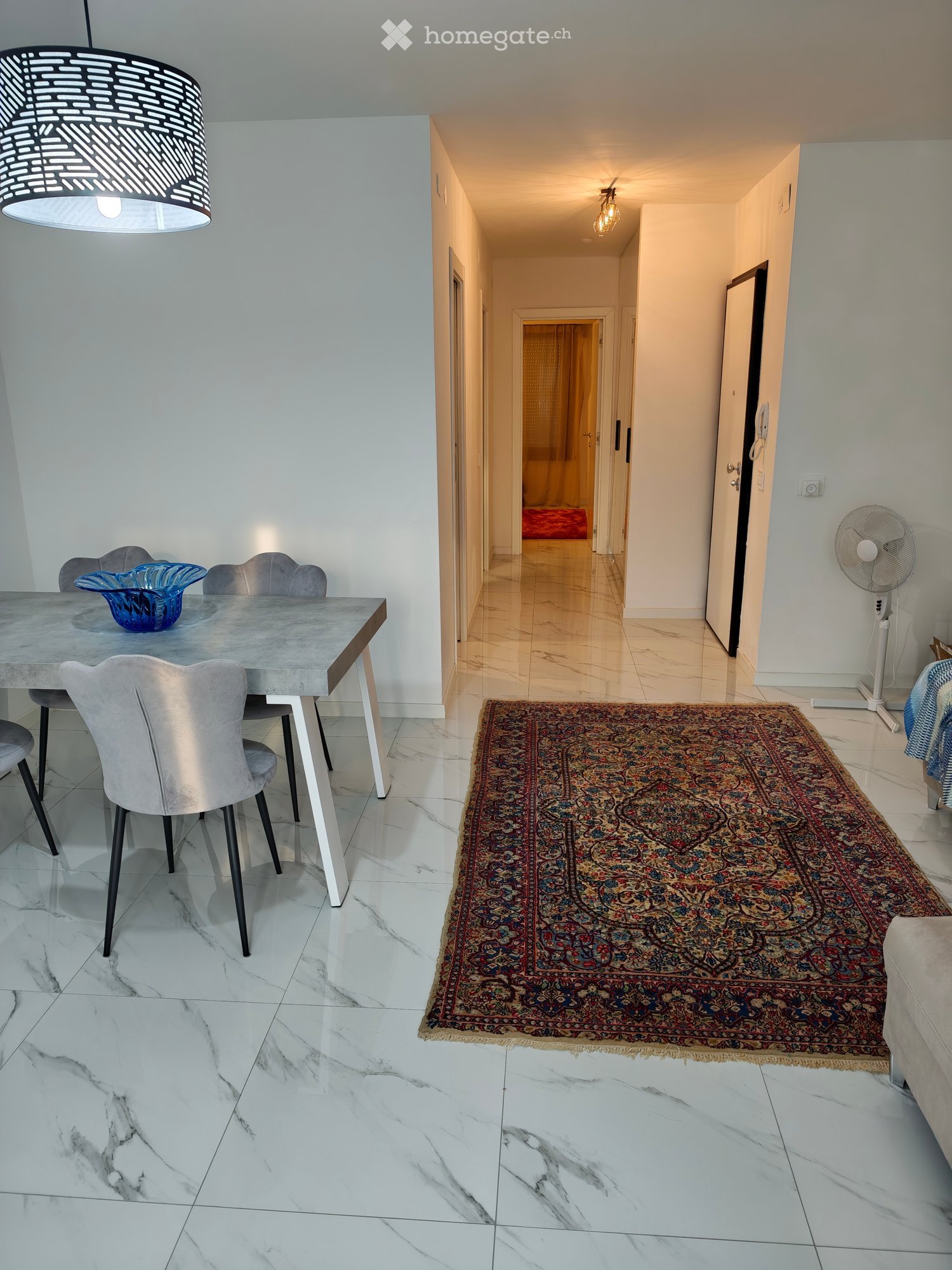 Modern hallway with marble flooring, a patterned rug, and a dining area visible to the left, featuring a grey table, chairs, and a decorative bowl.