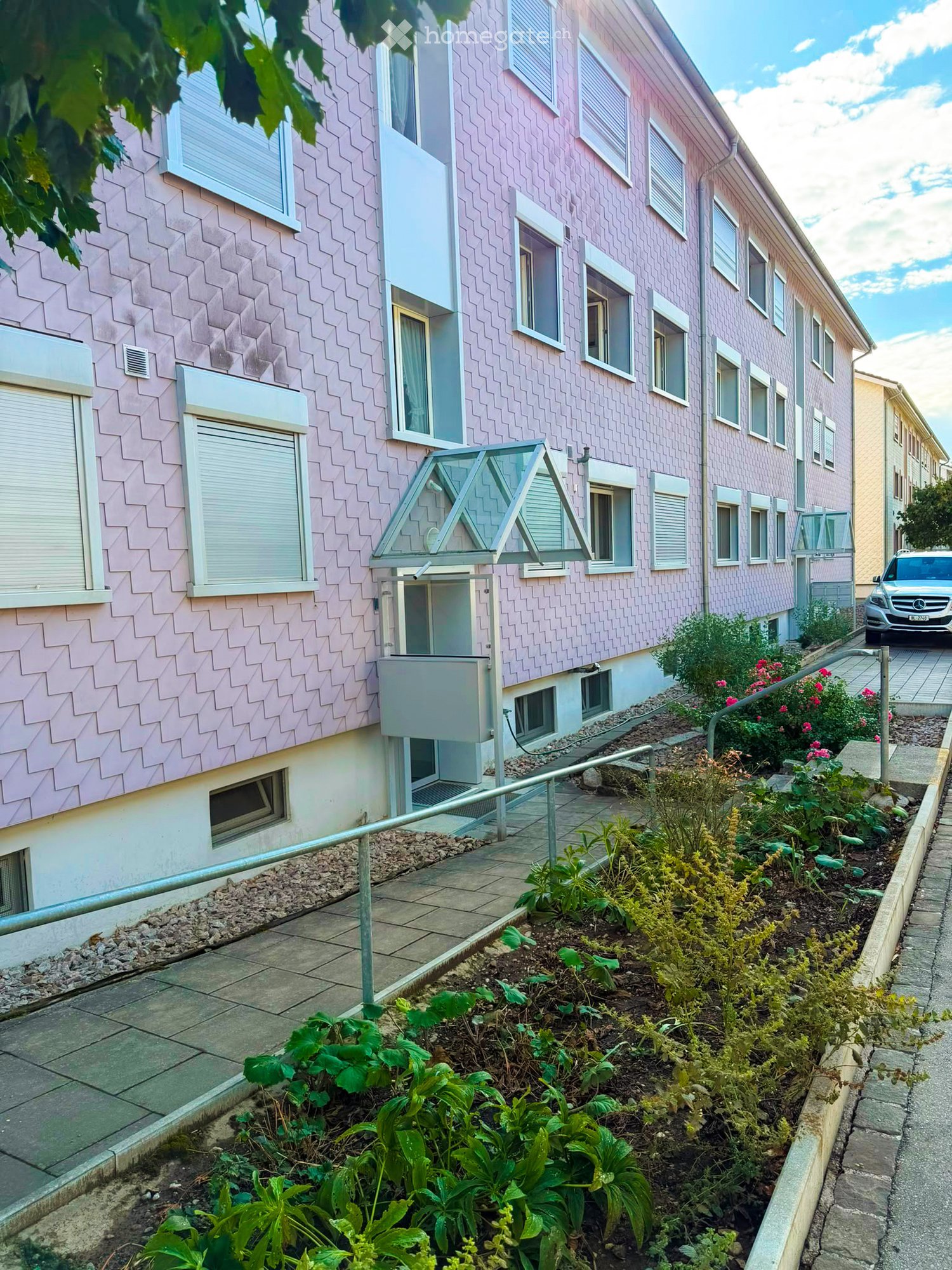 Pink residential building with multiple windows, a small garden area with plants and flowers, and a parked car in the background.
