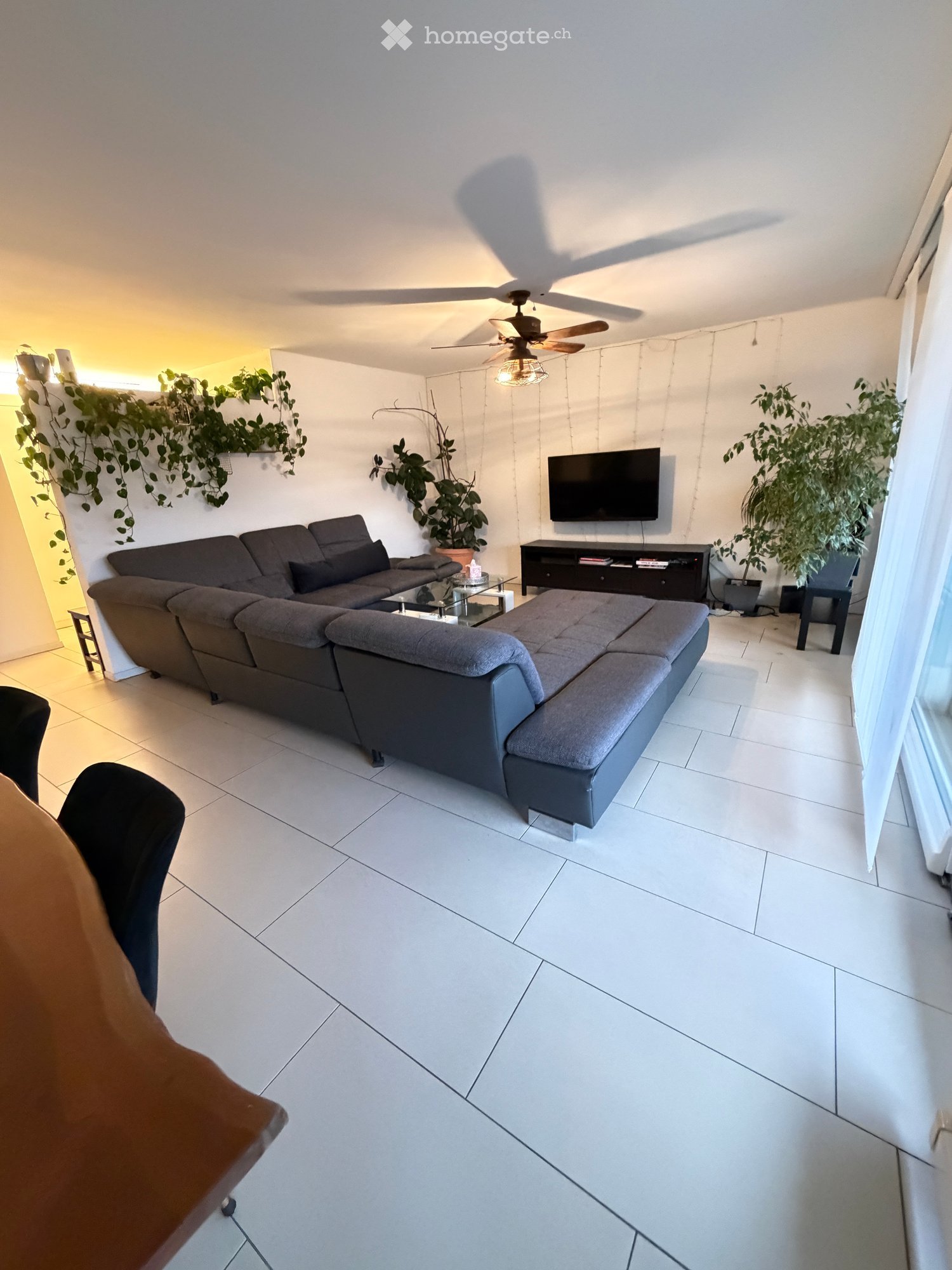Modern living room with white tiled flooring, grey sectional sofa, coffee table, TV stand, and plants.