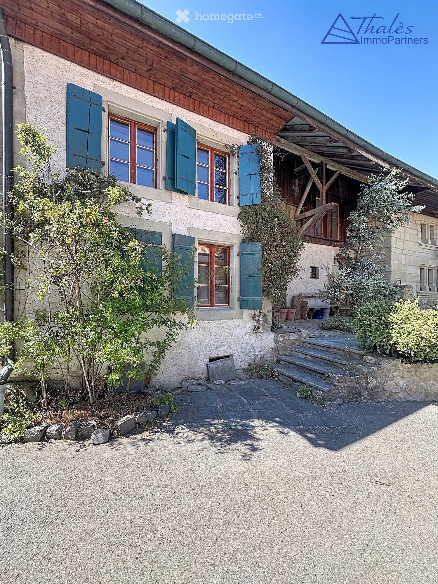 Two-story stone house with wooden shutters, a covered wooden porch, and a gravel driveway.
