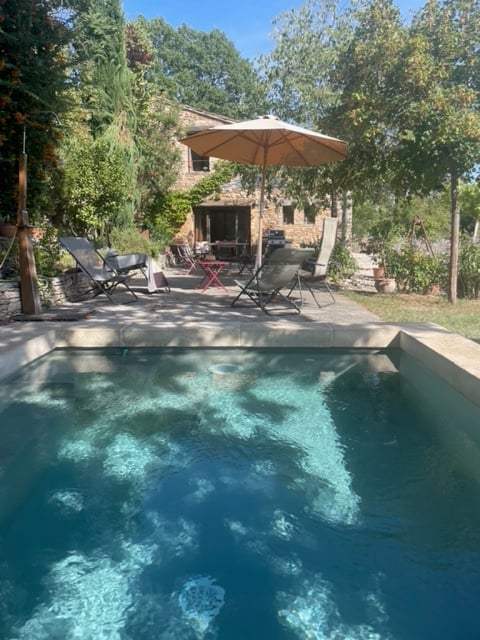 Outdoor patio area with a swimming pool, lounge chairs, umbrella, and a stone house in the background.