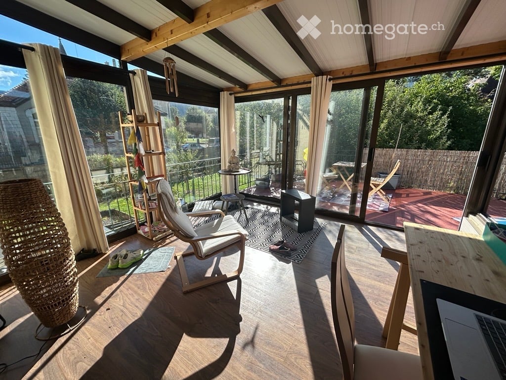 Bright, airy room with wooden flooring, large windows, and a balcony. Features a white chair, small table, stool, laptop, and decorative items.