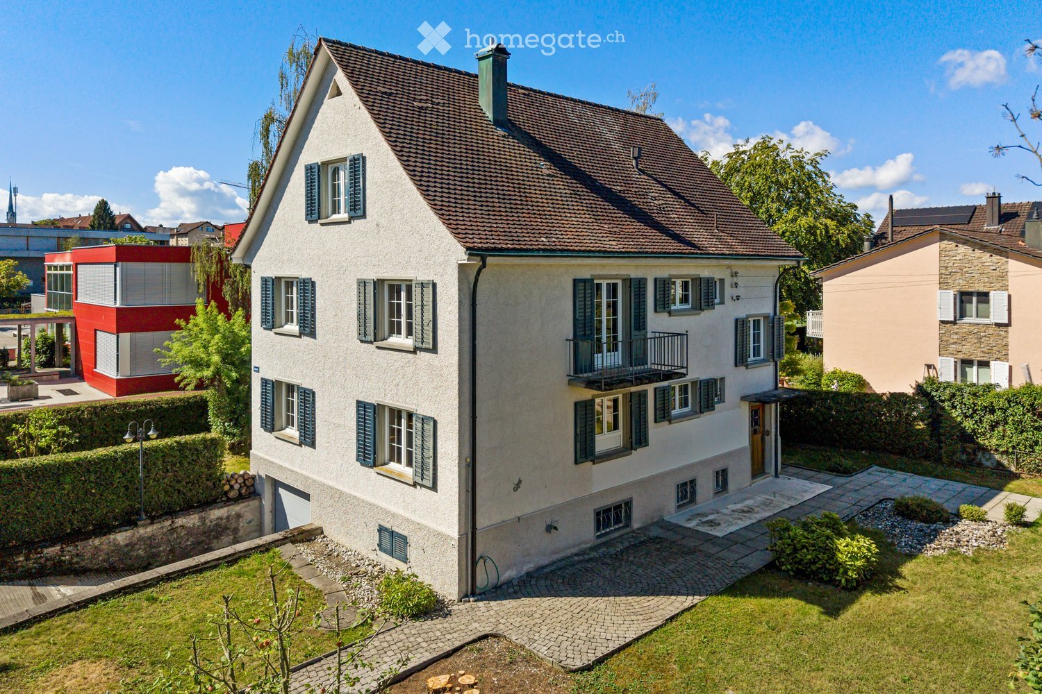 Two-story house with a gray facade, dark brown roof, multiple windows with shutters, and a small balcony on the upper floor. The house is surrounded by a well-maintained garden with a paved pathway leading to the entrance.