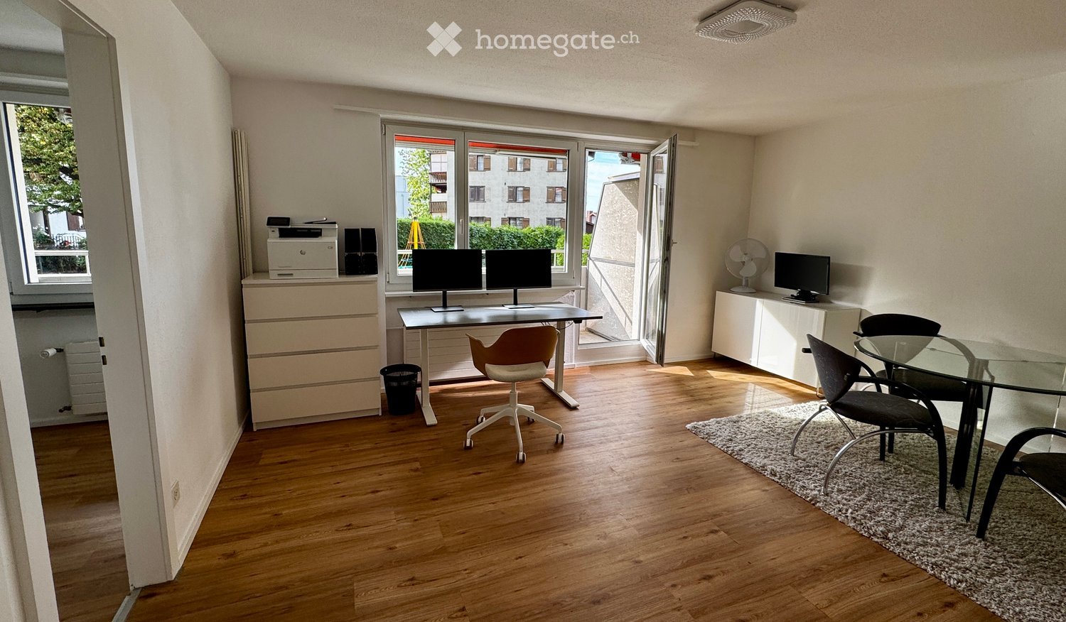 Bright, modern room with wooden flooring, large windows, and a balcony. Contains a desk with two monitors, a chair, a small table with chairs, a printer, and a fan.