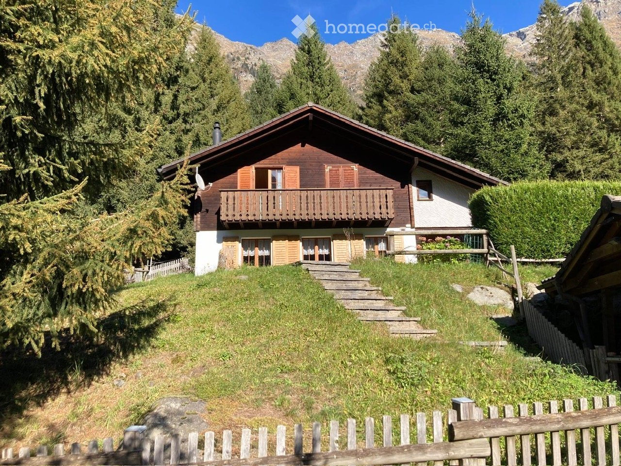 Two-story wooden house with a balcony, surrounded by trees and mountains.