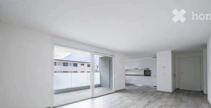 Bright, empty room with large sliding glass doors leading to a balcony, white walls, light wooden flooring, and a small adjacent kitchen area with white cabinets and appliances.