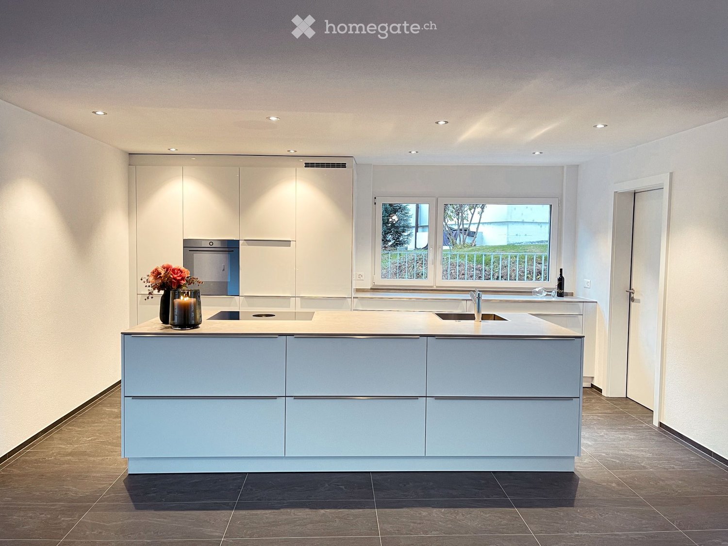 Modern kitchen with a central island, built-in cabinets, and a large window overlooking a green area. Features include a sink, faucet, candle, vase with flowers, and a bottle on the counter.