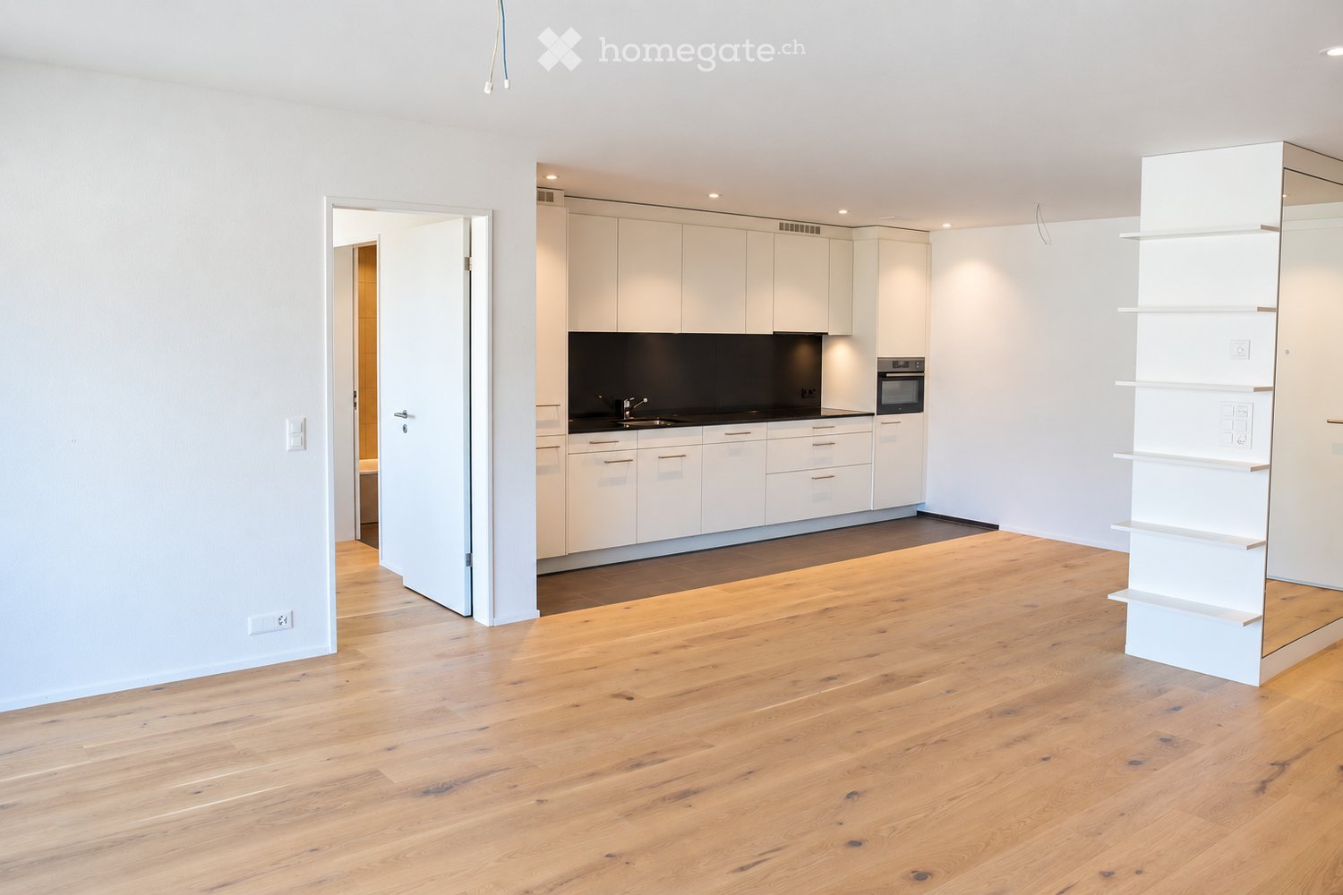 Open-plan living and kitchen area with light wooden flooring, white kitchen cabinets, a sink, and an oven.