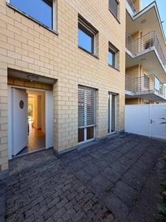 Modern multi-story apartment building with beige brick facade, large windows, and balconies. The ground floor features a paved entrance area with a visible doorway leading inside.