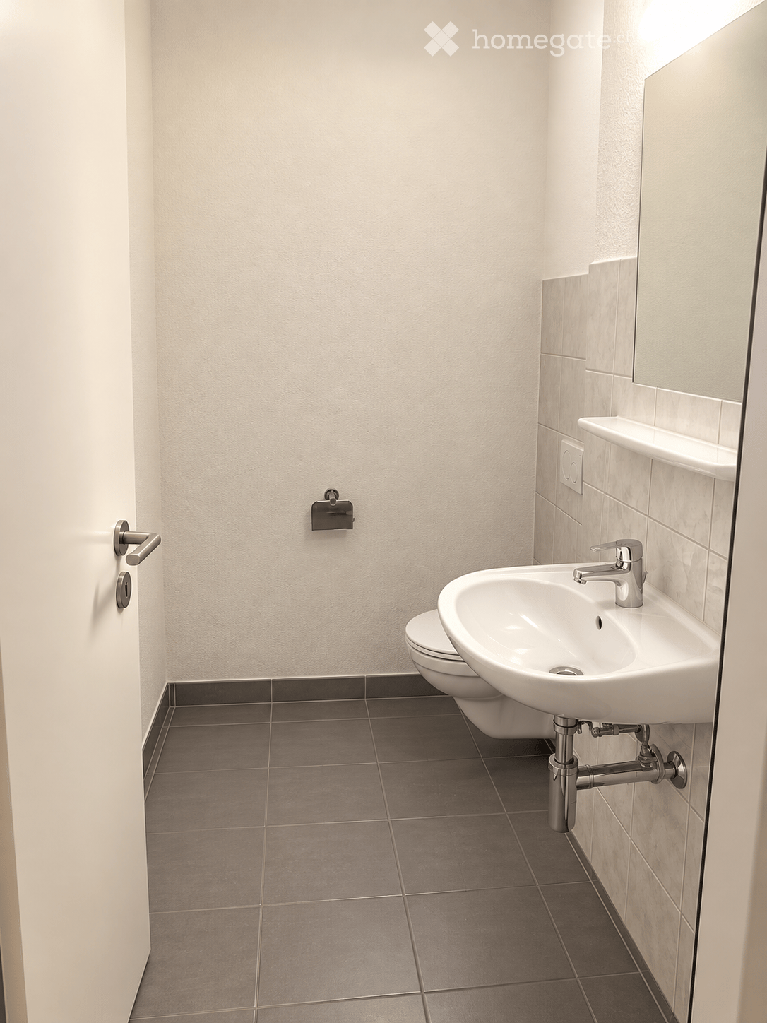 Modern bathroom with a white sink, chrome faucet, toilet, and a mirror above the sink. The walls are tiled in light beige, and the floor is covered with dark gray tiles.