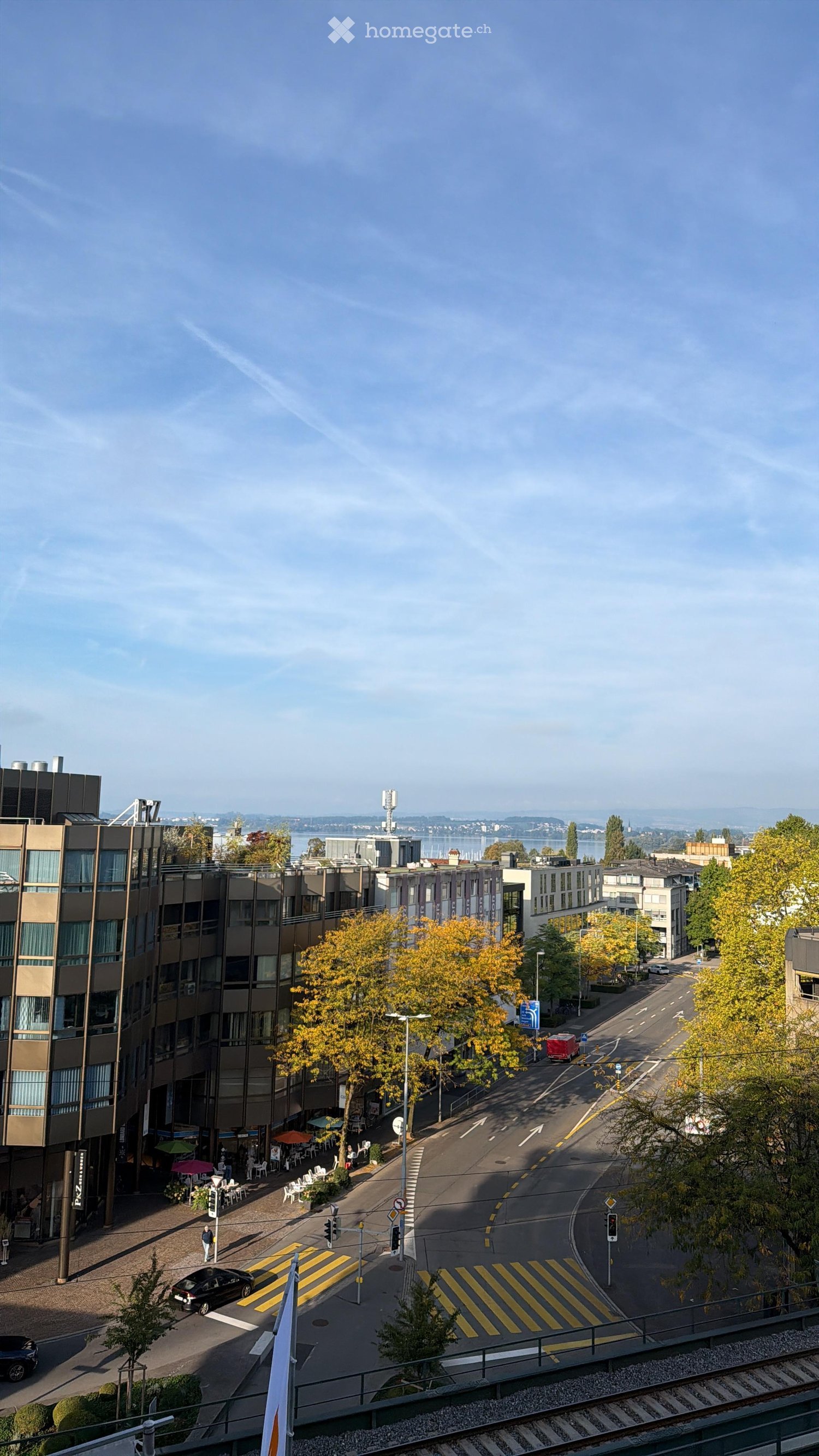 Aerial view of a city street with modern buildings, trees, traffic, and a pedestrian crossing.