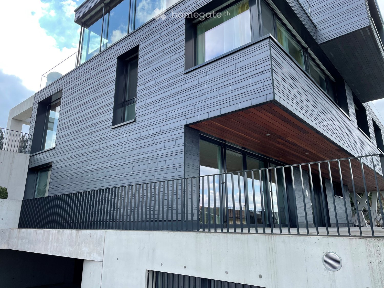 Modern multi-story building with grey brick facade, large windows, and a wooden balcony with metal railing.
