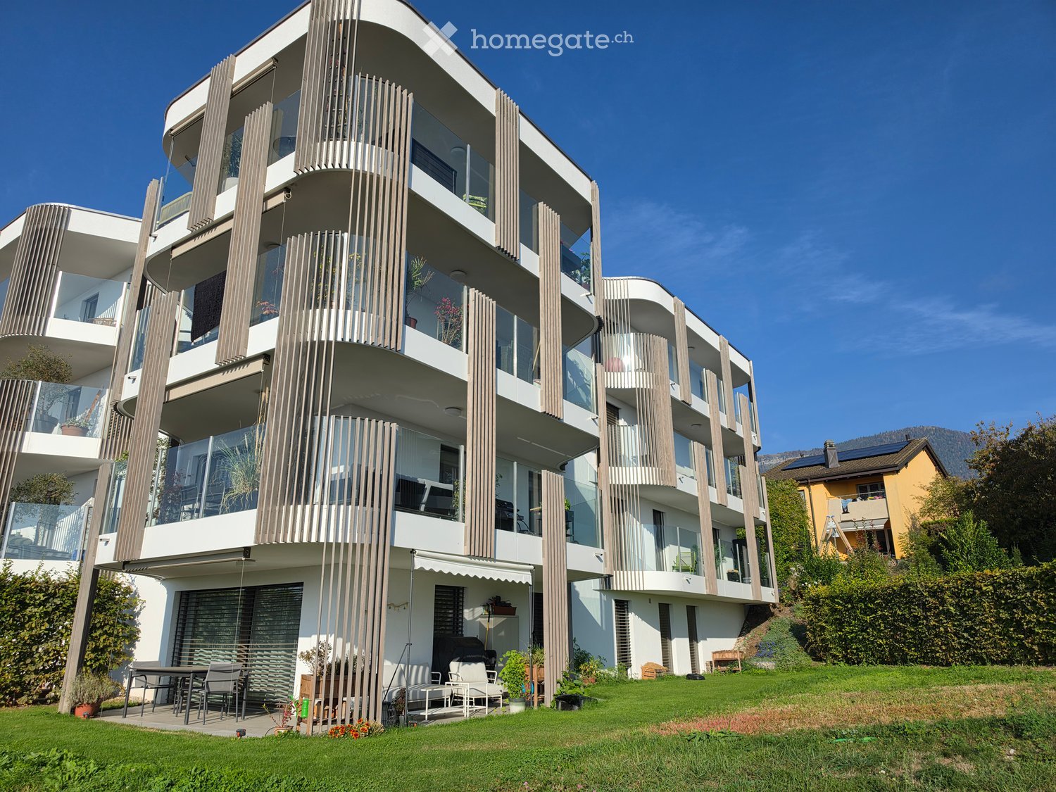 Modern multi-story building with balconies, large windows, and a raised ground floor, surrounded by a well-maintained lawn and outdoor seating area.