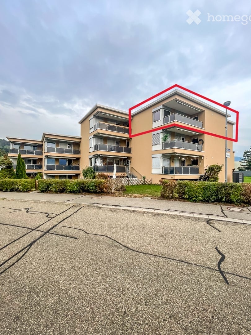Multi-story residential building with balconies and modern design, surrounded by greenery and a paved parking area.