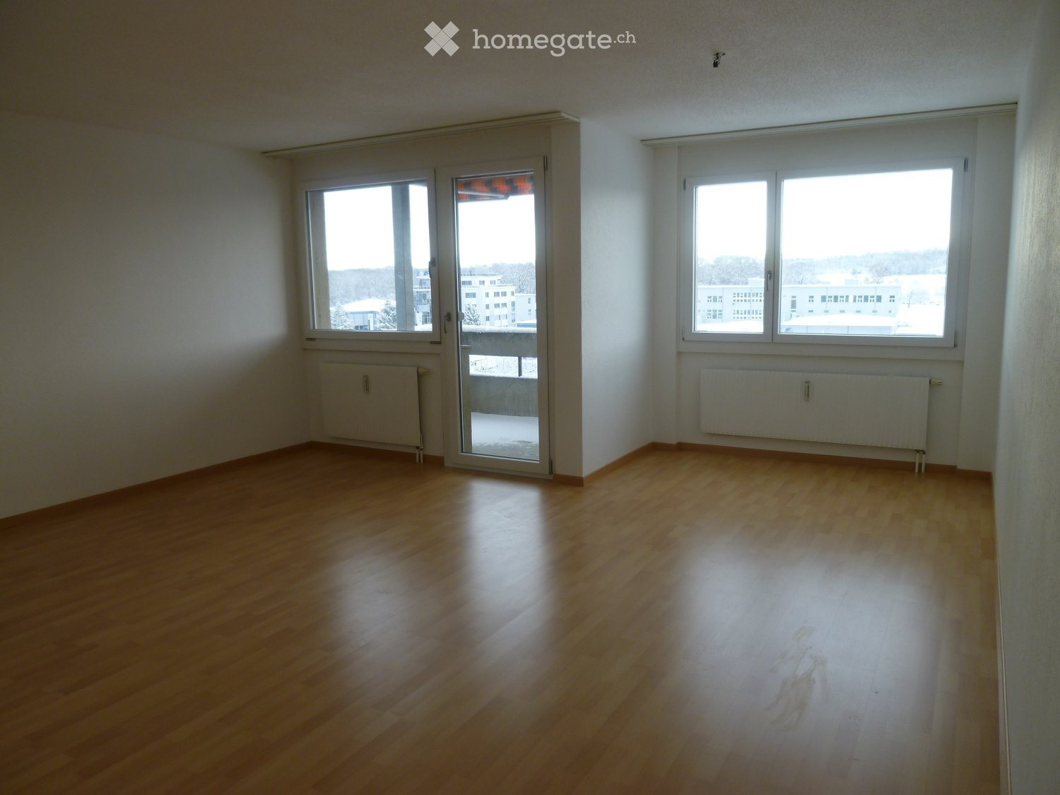 Empty room with wooden flooring, white walls, large windows, and a sliding glass door leading to a balcony.