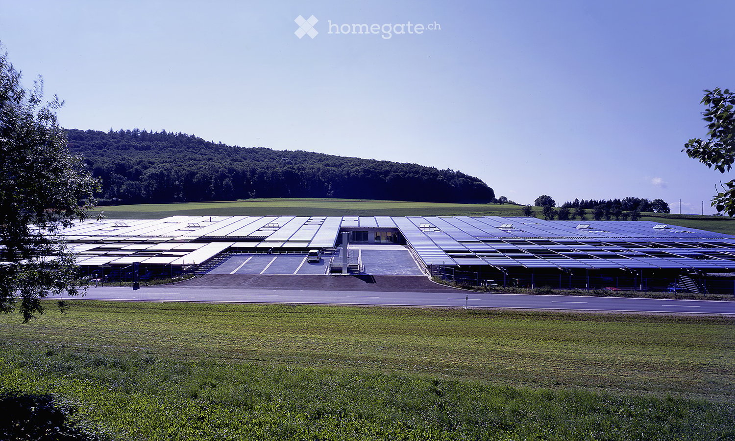 Large industrial building with solar panels on the roof, surrounded by open fields and a parking area.