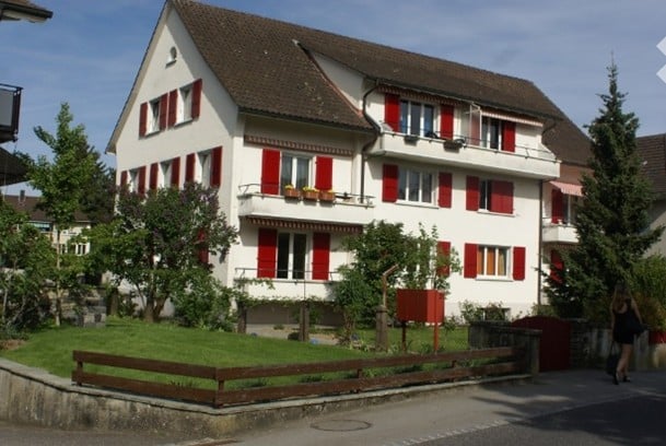 A two-story house with white walls, red shutters, and a brown roof. The house features multiple windows, balconies, and is surrounded by greenery and a wooden fence.