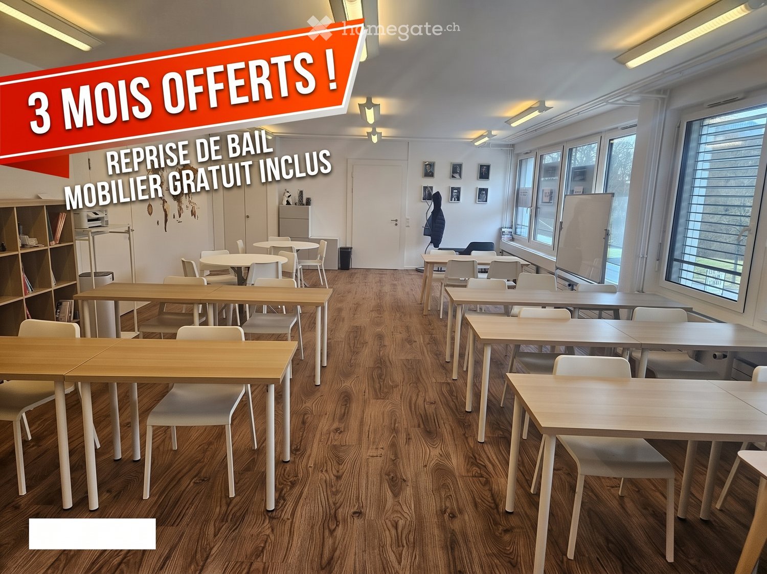 Bright, modern room with wooden flooring, multiple tables and chairs arranged for a collaborative workspace, shelves, a refrigerator, a printer, and a whiteboard.