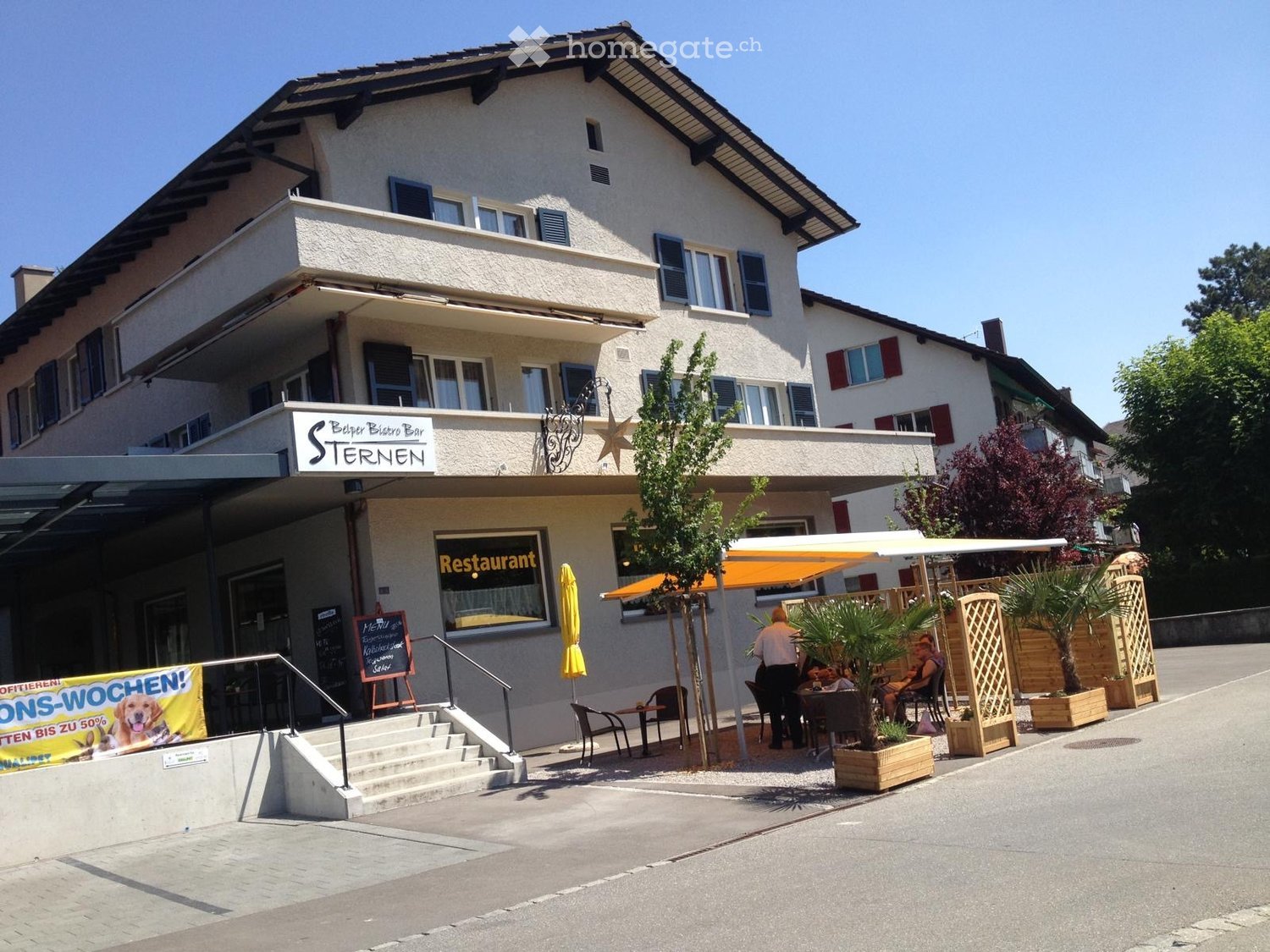 Two-story building with a restaurant sign, outdoor seating area, and a small garden with potted plants.