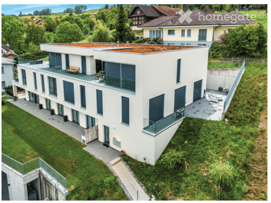 Modern two-story house with white exterior, large windows, balconies, and a flat roof. The house is surrounded by a well-maintained garden with greenery and a pathway leading to the entrance.