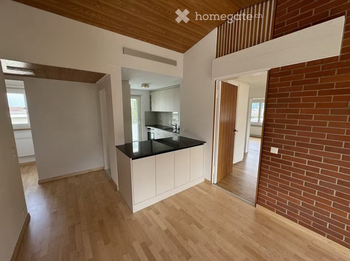 Open-plan kitchen area with white cabinets, black countertop, and a central island. Wooden flooring and exposed wooden ceiling beams.