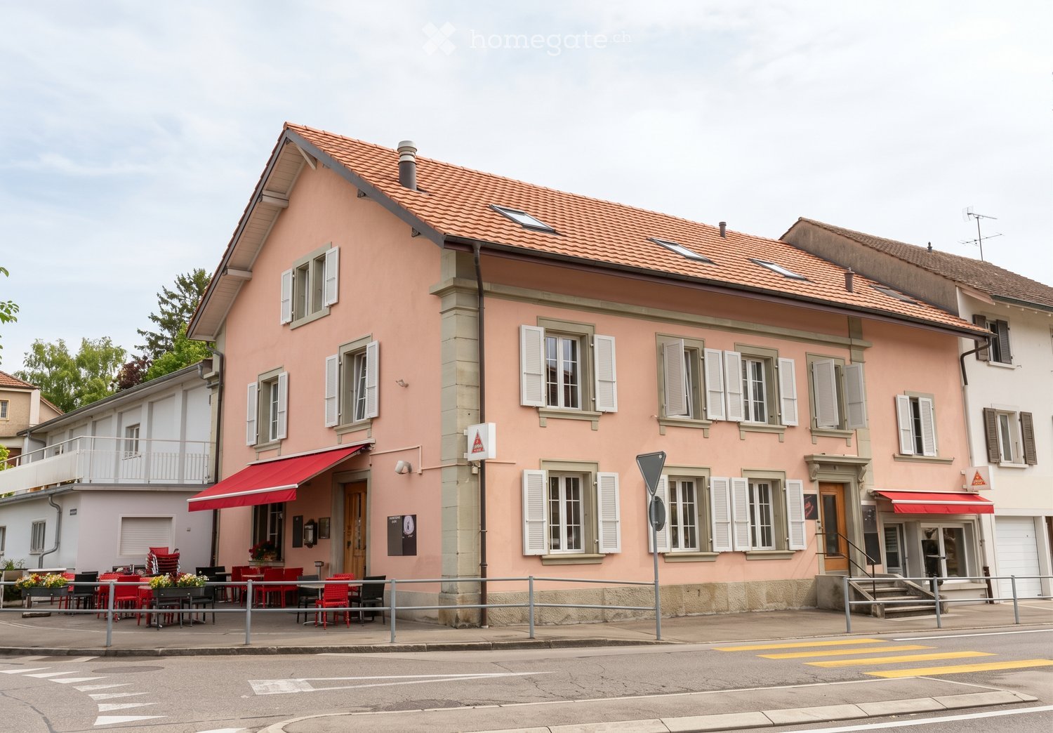 A two-story pink building with white shutters, red awnings, and outdoor seating area with red chairs and tables.