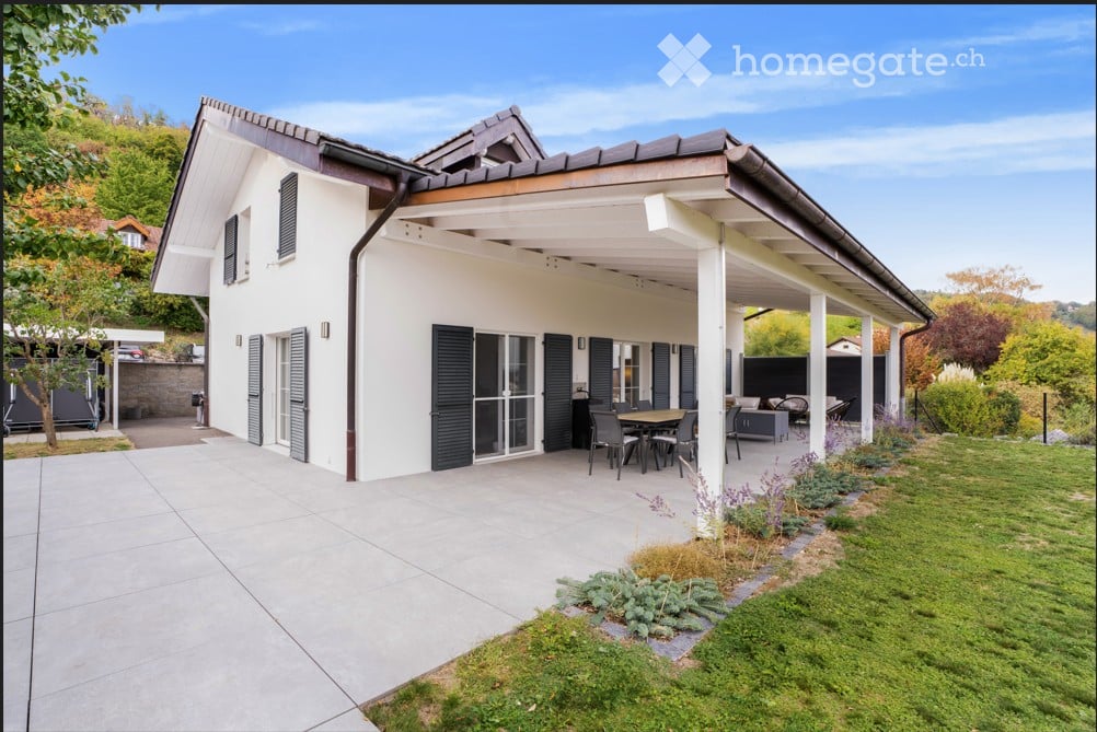 Modern two-story house with white exterior, dark shutters, and a covered patio area with outdoor furniture.