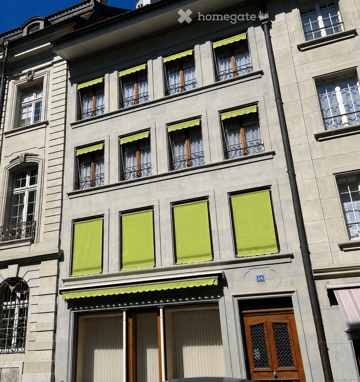 A multi-story building with light-colored stone facade, multiple windows with green awnings, and ornate metal railings on balconies.