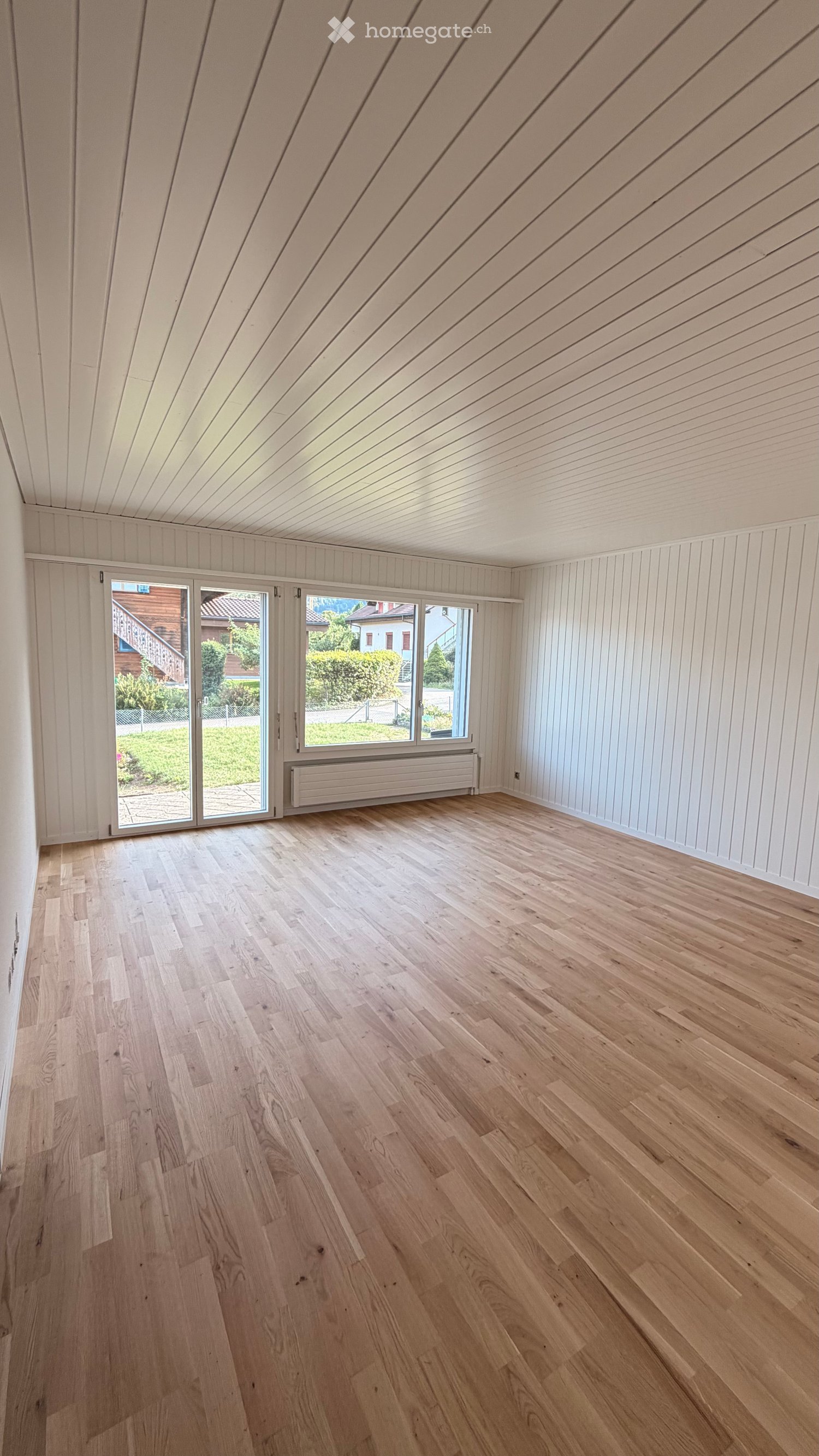 Empty room with light wooden flooring, white paneled walls, and a slanted wooden ceiling. Large windows and sliding doors lead to an outdoor area.